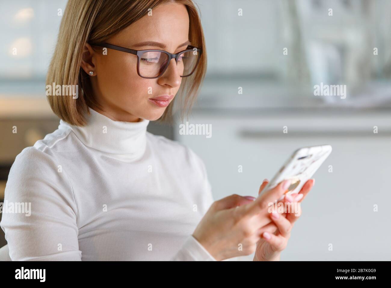 Donna d'affari seria indossa il maglione e gli occhiali turleneck bianco utilizzando smartphone moderno in custodia di marmo, navigare in internet, buyng merci online, sfocato Foto Stock