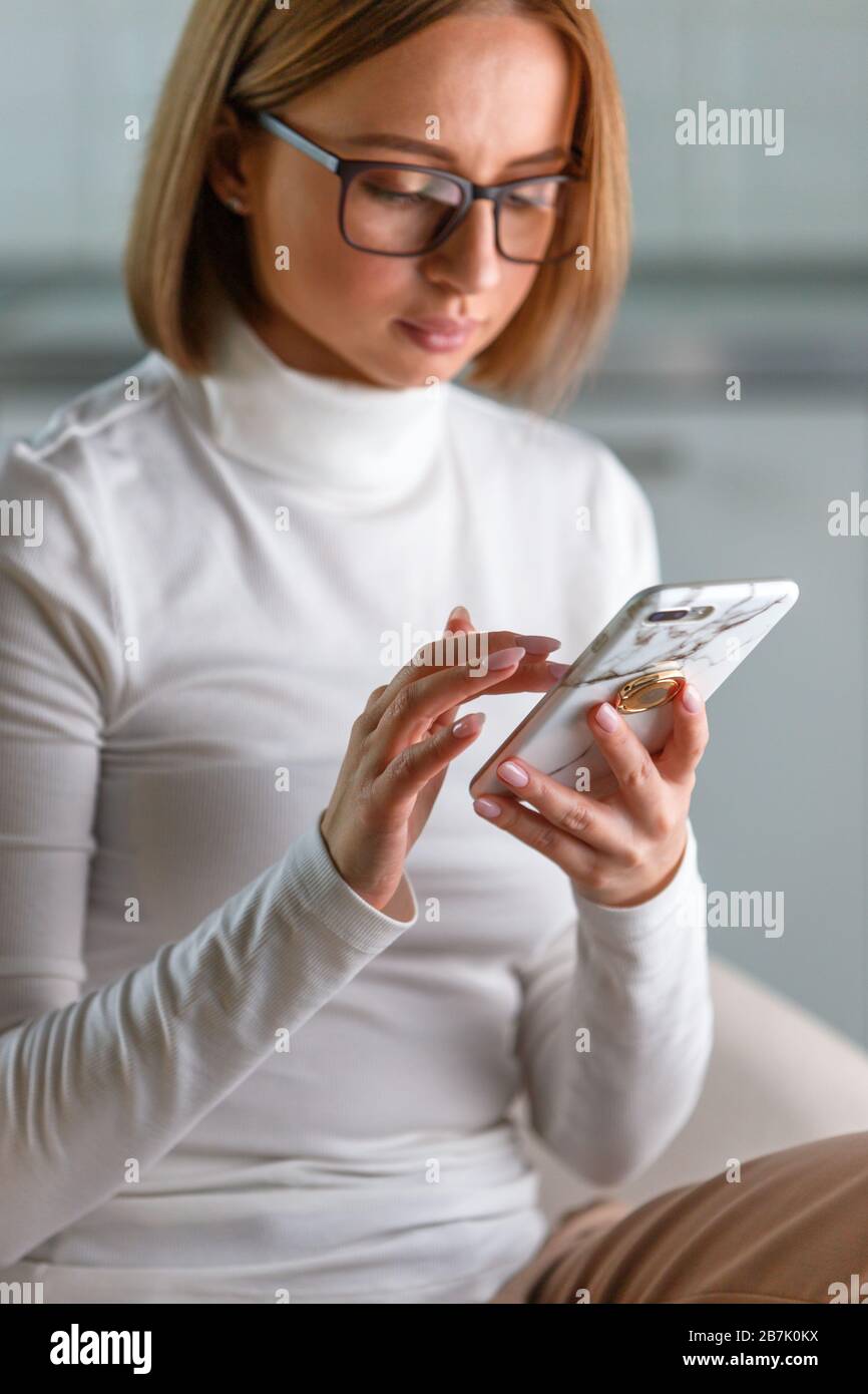 Donna d'affari seria indossa il maglione e gli occhiali turleneck bianchi in custodia di marmo, navigare in internet, acquistare merci online, sfondo sfocato. Rete sociale Foto Stock