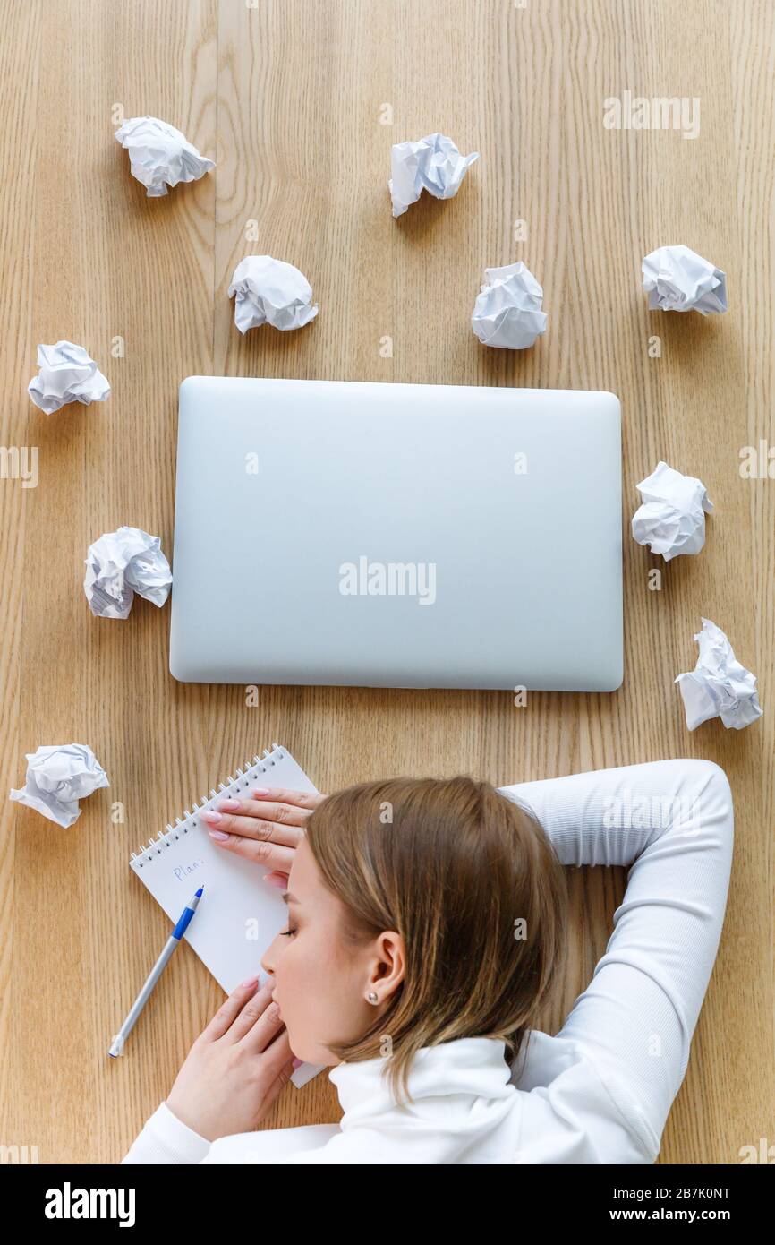 Donna stanca freelancer che dorme e riposa la testa sul notebook sul tavolo, scrivendo su blocco note, palle di carta sbriciolate e laptop intorno, vista dall'alto Foto Stock