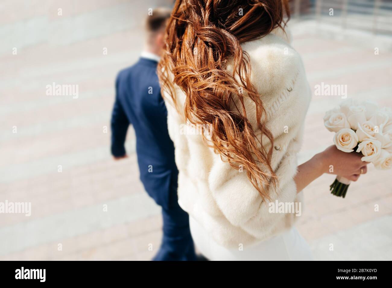 coppia di amanti del matrimonio alla moda in strada tornano. La sposa in un leggero cappotto di pelliccia con lunghi capelli ricci scuri contiene un bouquet bianco di rose. Foto Stock