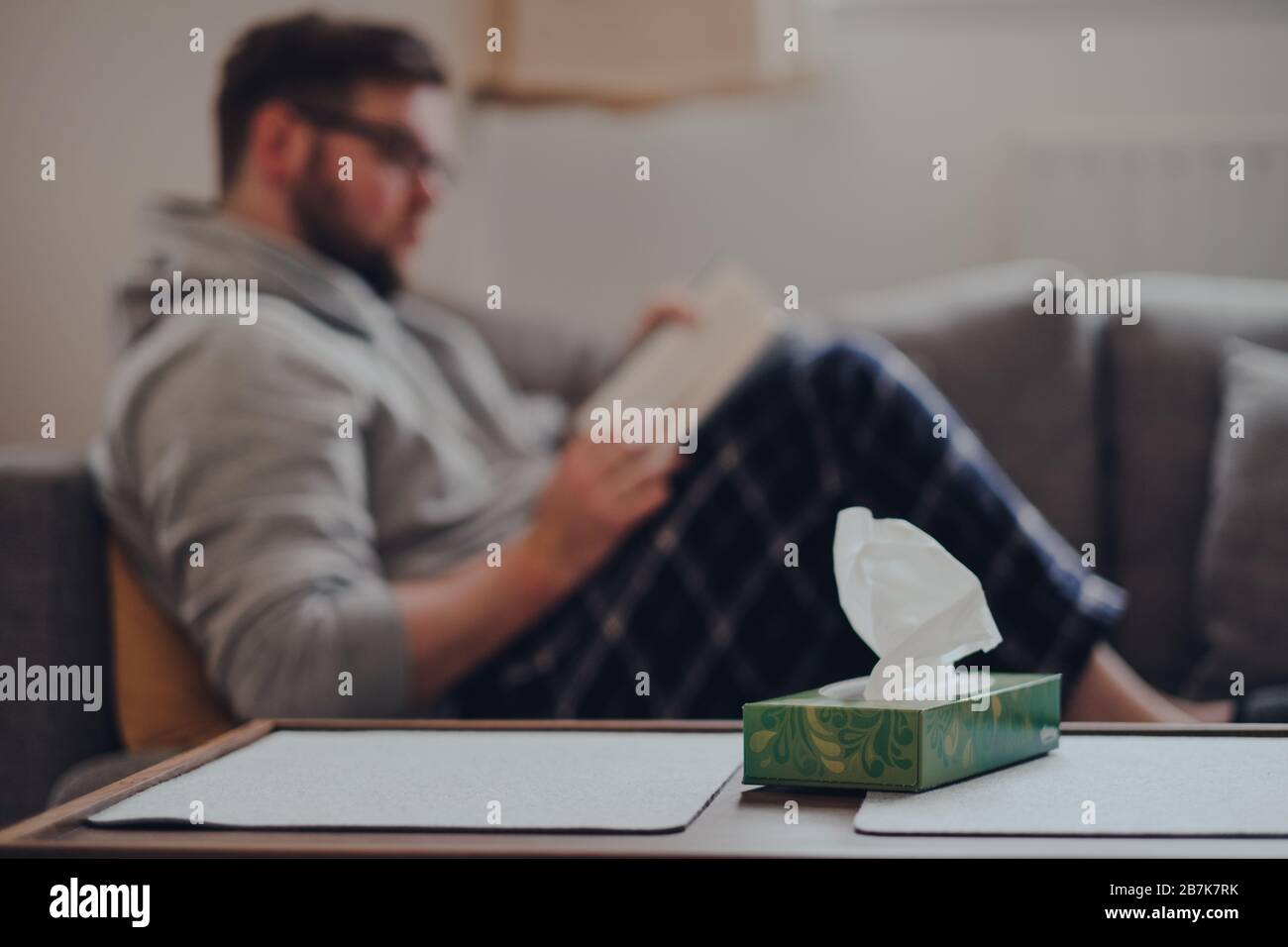 Scatola di tessuti sul tavolo, uomo sullo sfondo seduto su un divano, lettura di un libro. Concetto di malato a casa. Foto Stock