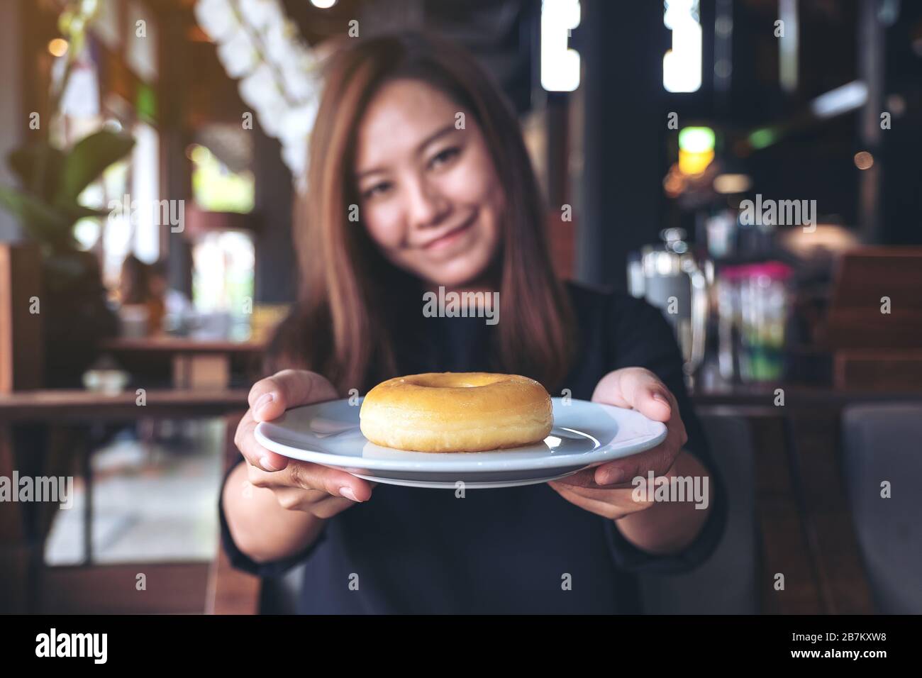 Una bella donna asiatica che tiene e mostra un piatto di ciambella con sentirsi felice e buon stile di vita nel moderno caffè Foto Stock