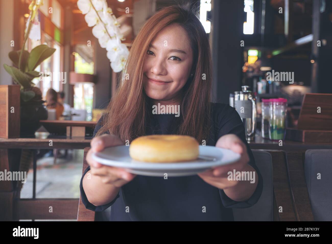 Una bella donna asiatica che tiene e mostra un piatto di ciambella con sentirsi felice e buon stile di vita nel moderno caffè Foto Stock