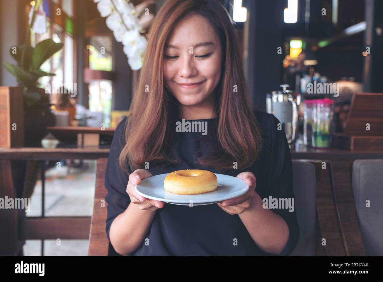 Una bella donna asiatica che tiene e mostra un piatto di ciambella con sentirsi felice e buon stile di vita nel moderno caffè Foto Stock