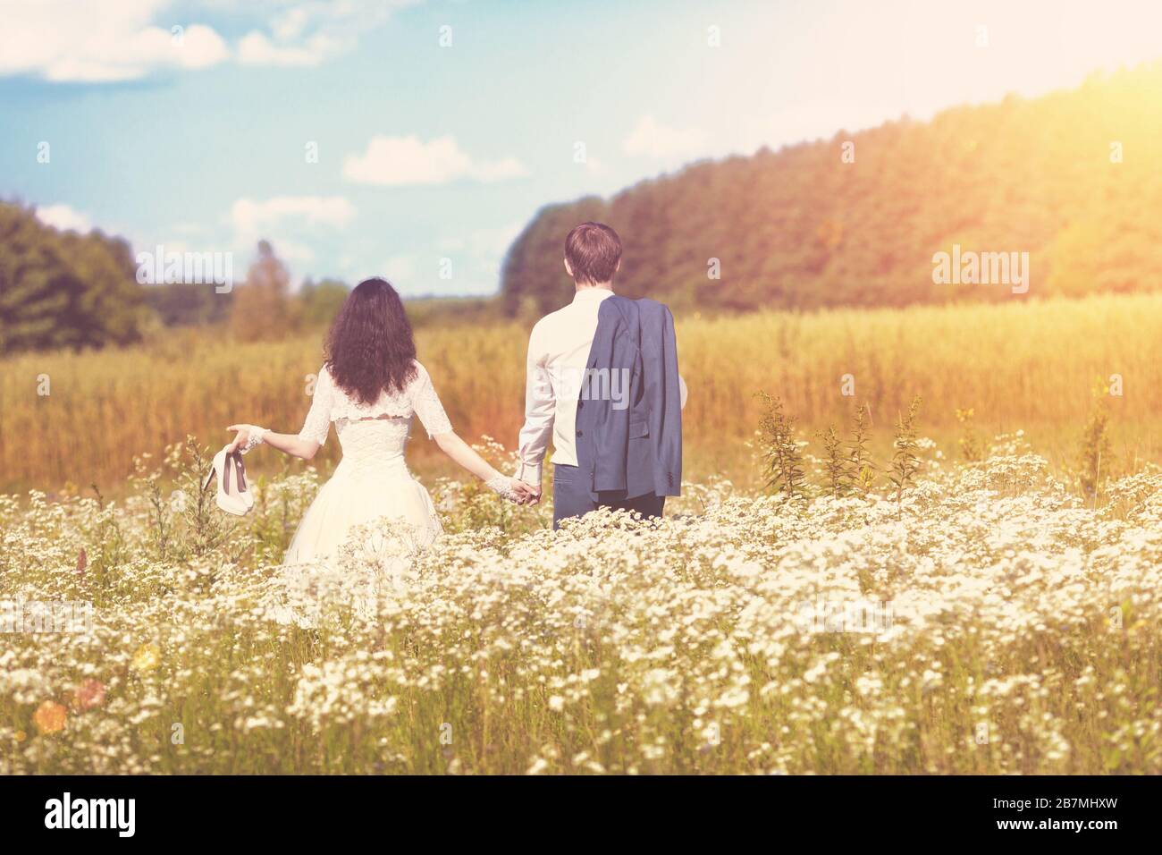 Una giovane coppia felice di nozze che cammina in un campo fiorito e tiene le mani su una giornata estiva soleggiata. Una donna tiene le scarpe nelle sue mani, un uomo tiene un jac Foto Stock