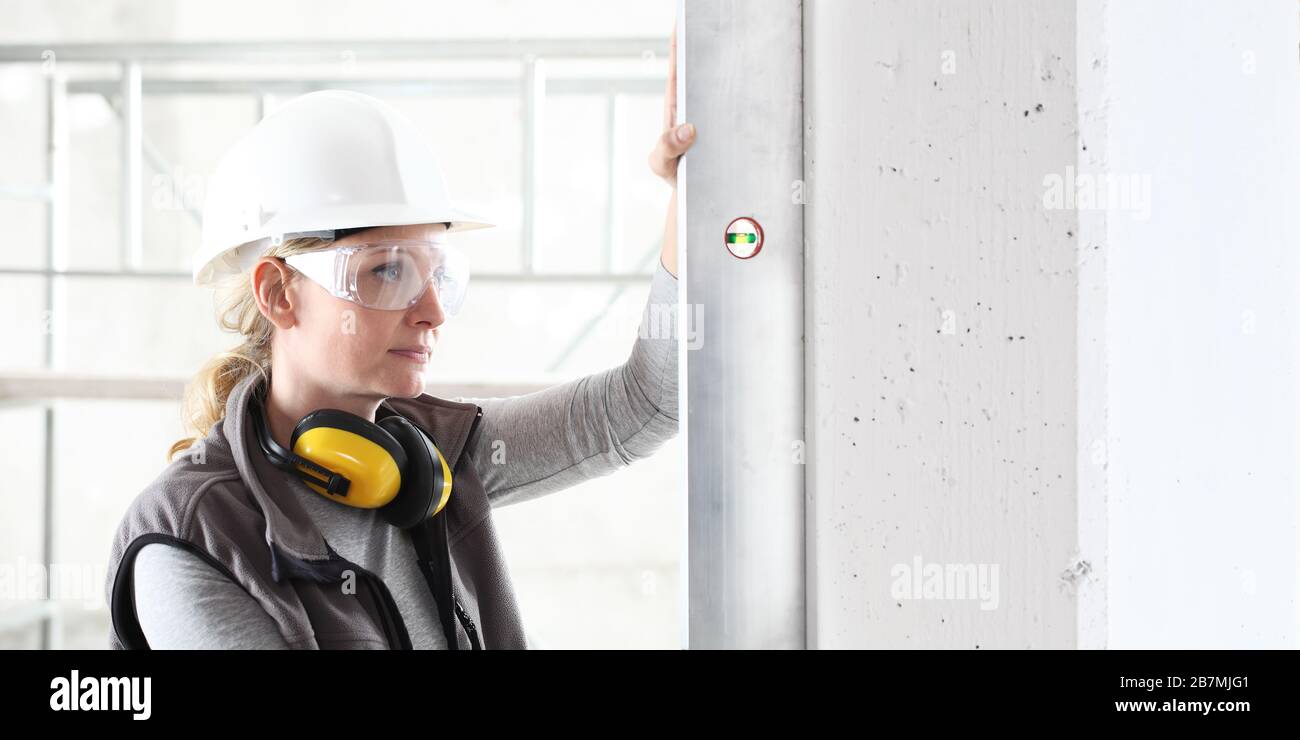 donna lavoratore guarda la livella dello spirito controlla la parete con elmetto, occhiali e cuffie di protezione dell'orecchio, parete bianca con spazio di copia Foto Stock
