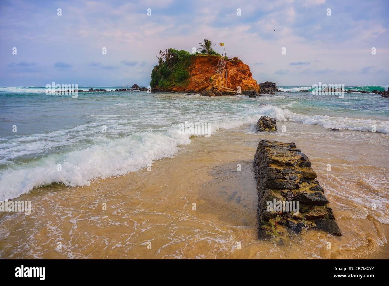 Tempo tempestoso sulla costa dell'Oceano Indiano in Sri Lanka. Nuvole di sole e piccole increspature dell'oceano. Il muro rovinato si stacca dalla sabbia del mare. Foto Stock
