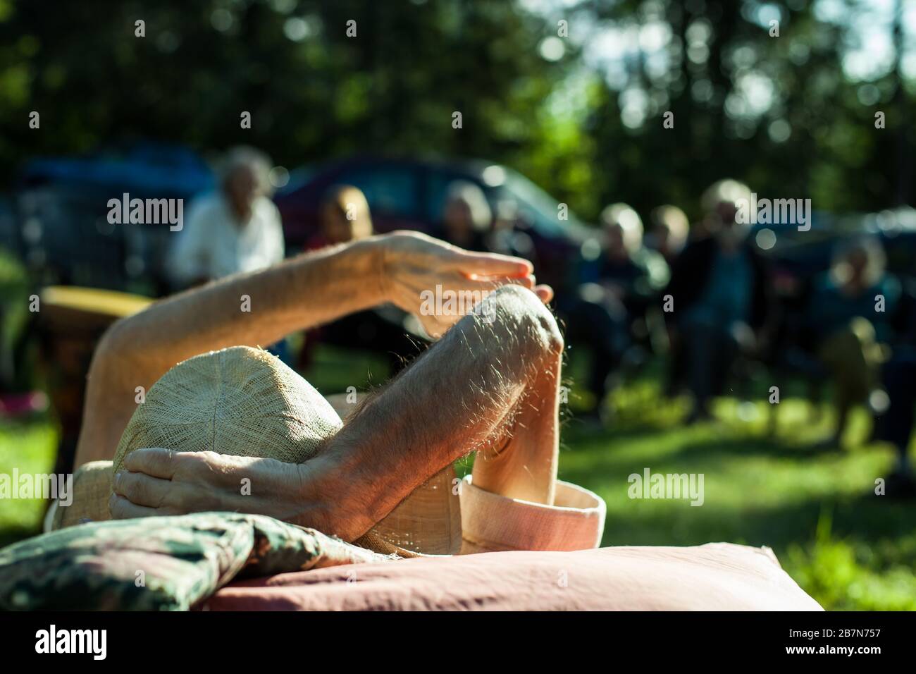 Un primo piano di fuoco selettivo girato da dietro un vecchio uomo morente è alla sua festa di addio, con i membri della famiglia sfocati in background, all'aperto al sole. Foto Stock