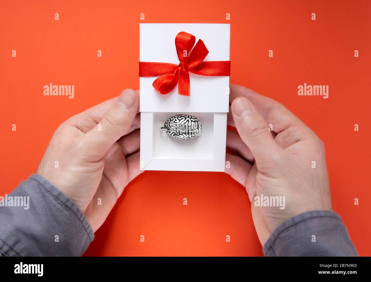 Mani maschili che tengono una scatola regalo bianca aperta a metà con un modello in metallo di un cervello umano all'interno e un arco rosso con un nastro sulla copertura. Foto Stock