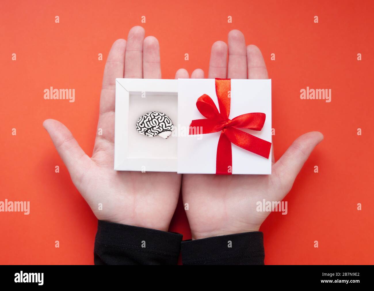 Mani femminili che tengono una scatola regalo bianca aperta con un modello in acciaio di un cervello umano all'interno e un nastro rosso sulla copertura. Concetto creativo attuale. Foto Stock