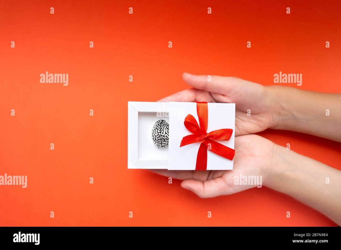 Mani femminili che tengono una scatola regalo bianca con un modello metallico di cervello umano all'interno e un arco rosso sulla copertura. Concetto creativo attuale. Foto Stock