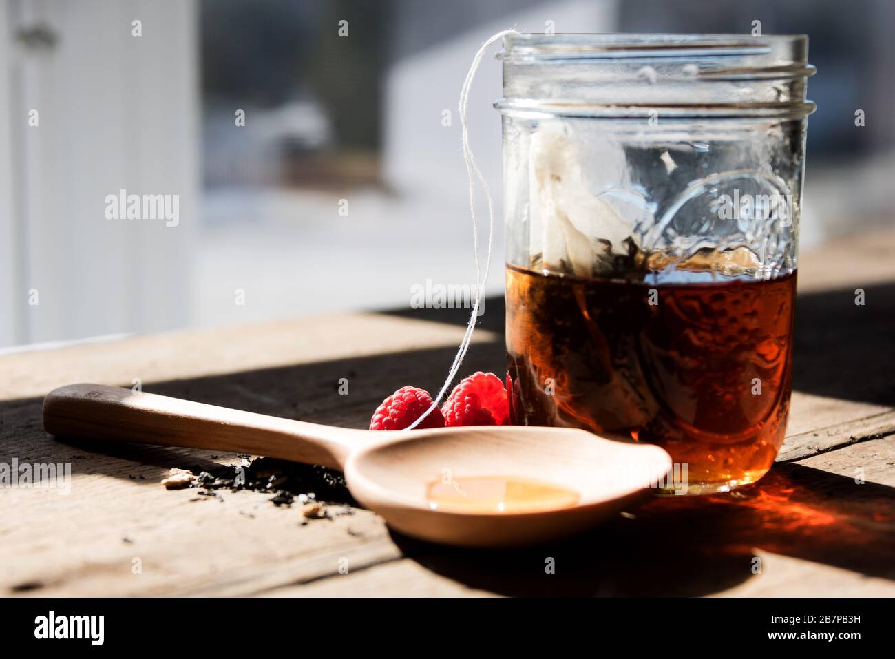 Sacchetto di tè fatto in casa che si stacca in un vaso di muratura su un tavolo di legno davanti ad una finestra, con tè, lamponi e un cucchiaio di legno riempito di miele. Foto Stock