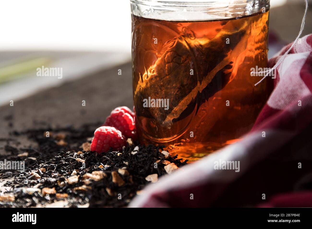 Sacchetto di tè fatto in casa che si macina in un vaso di mason, con tè e lamponi versati davanti, e un panno rosso-e-bianco controllato dietro. Foto Stock