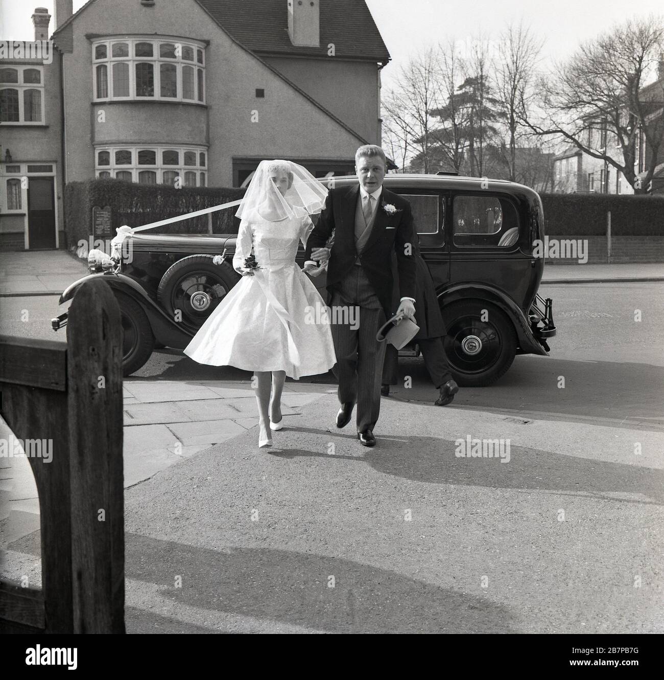 1960, storico, fuori in una strada suburbana, una macchina d'epoca parcheggiata strada, come una sposa arriva per il suo matrimonio, sud-est di Londra, Inghilterra, Regno Unito. Foto Stock