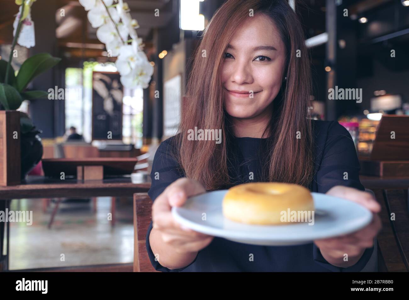 Una bella donna asiatica che tiene e mostra un piatto di ciambella con sentirsi felice e buon stile di vita nel moderno caffè Foto Stock