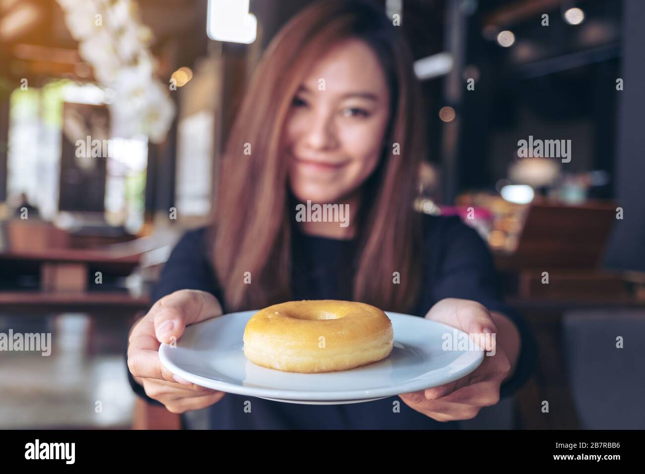 Una bella donna asiatica che tiene e mostra un piatto di ciambella con sentirsi felice e buon stile di vita nel moderno caffè Foto Stock