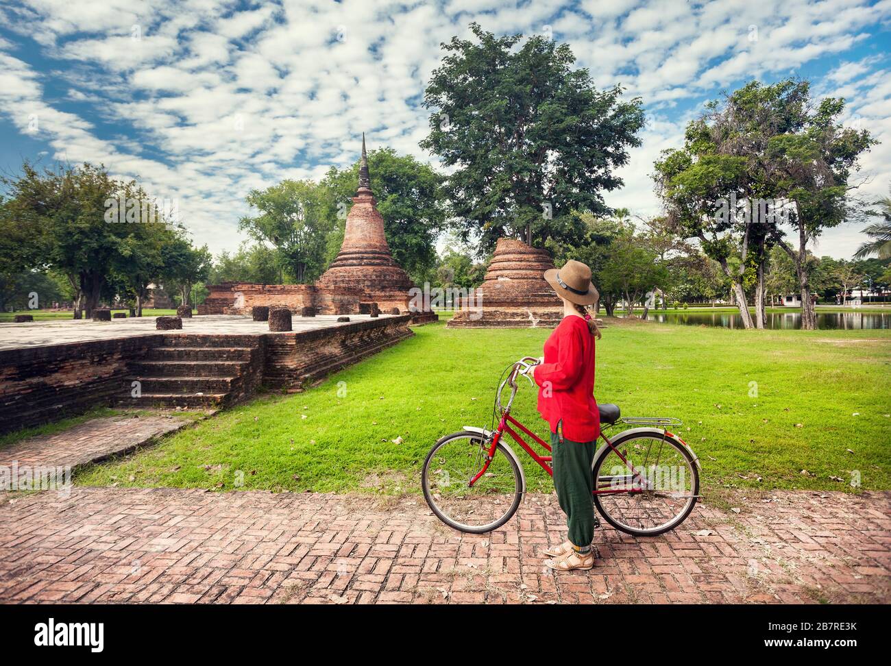 Donna in maglietta rossa con bicicletta guardando vecchi rovinato tempio buddista in Sukhothai historical park, Thailandia Foto Stock