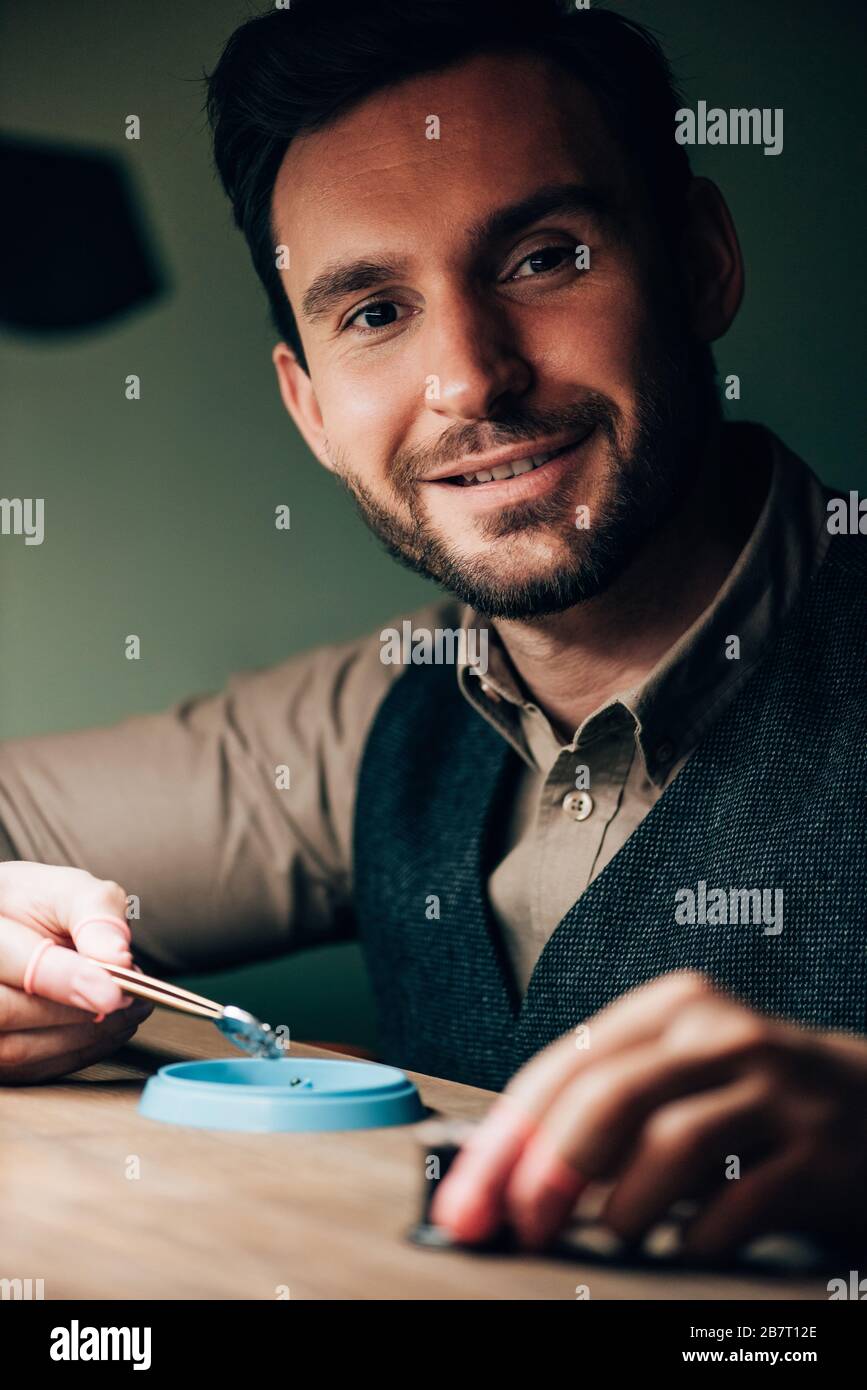 Bell'orologiaio sorridente alla macchina fotografica mentre lavora con le parti dell'orologio al tavolo Foto Stock