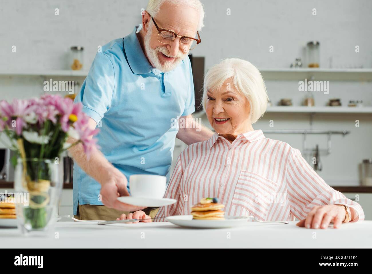 L'attenzione selettiva dell'uomo anziano che mette il caffè sul tavolo da una moglie sorridente durante la colazione Foto Stock