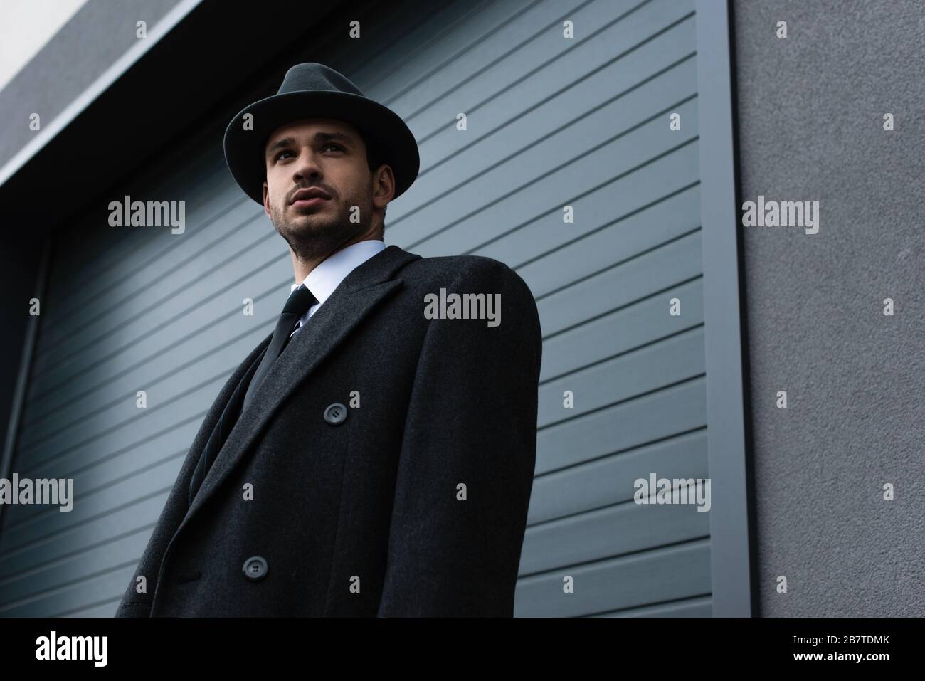 Vista a basso angolo del mafioso in cappotto nero e cappello di feltro  vicino al muro sulla strada Foto stock - Alamy