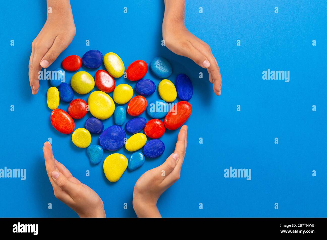 Concetto del giorno di consapevolezza di autismo del mondo. Mani per bambini con cuore fatte di pietre di colore rosso giallo blu su sfondo blu chiaro Foto Stock