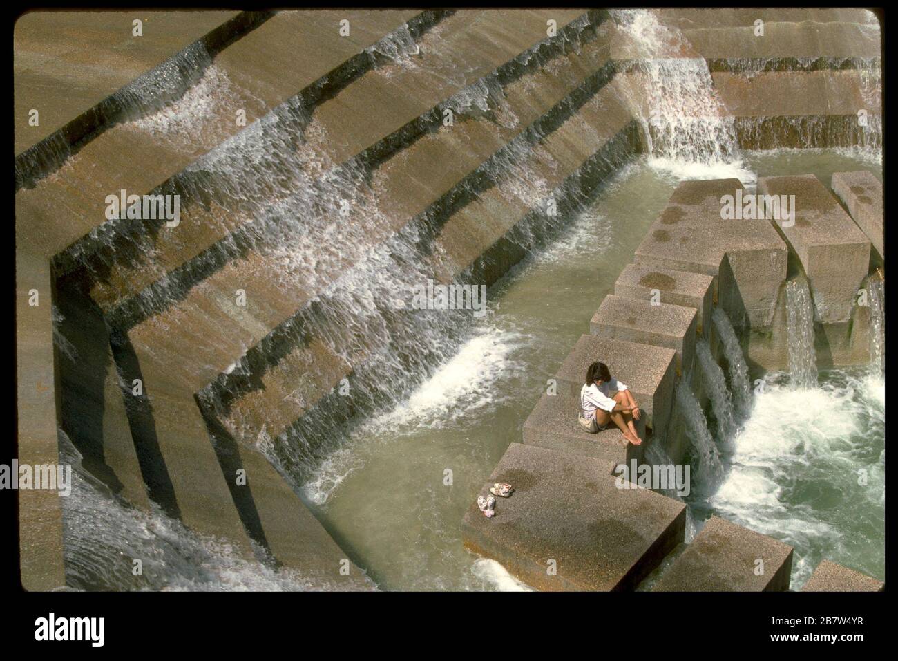 Fort Worth Texas USA: La donna gode di viste, suoni e sensazione di cascata d'acqua, mentre si siede su un blocco di cemento nella piscina attiva dei giardini acquatici di Fort Worth nel centro città. ©Bob Daemmrich/ Foto Stock