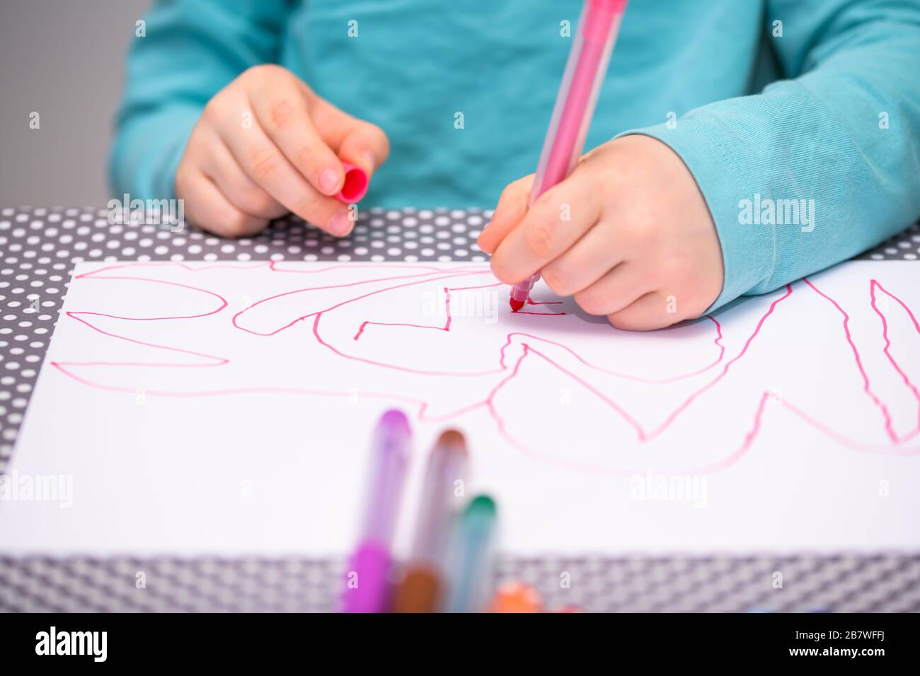 Primo piano di un ragazzo di cinque anni che gioca e disegna con una penna di colore rosa su una carta bianca posta su un tavolo con puntini bianchi. Foto Stock
