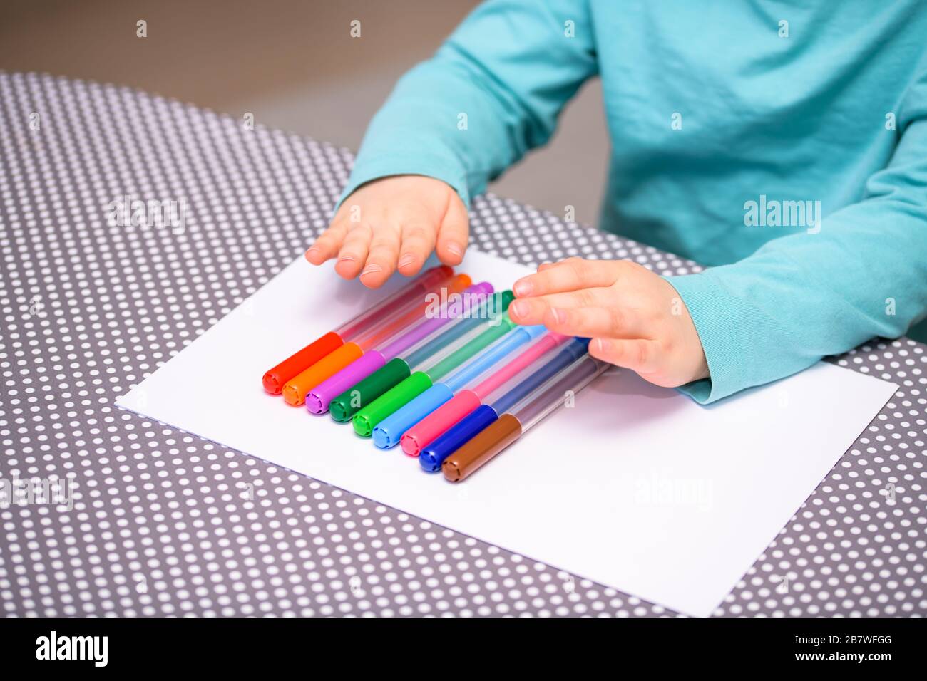 Primo piano di un ragazzo di cinque anni che gioca e allineano le penne a colori su una carta bianca posta su un tavolo con puntini bianchi. Foto Stock