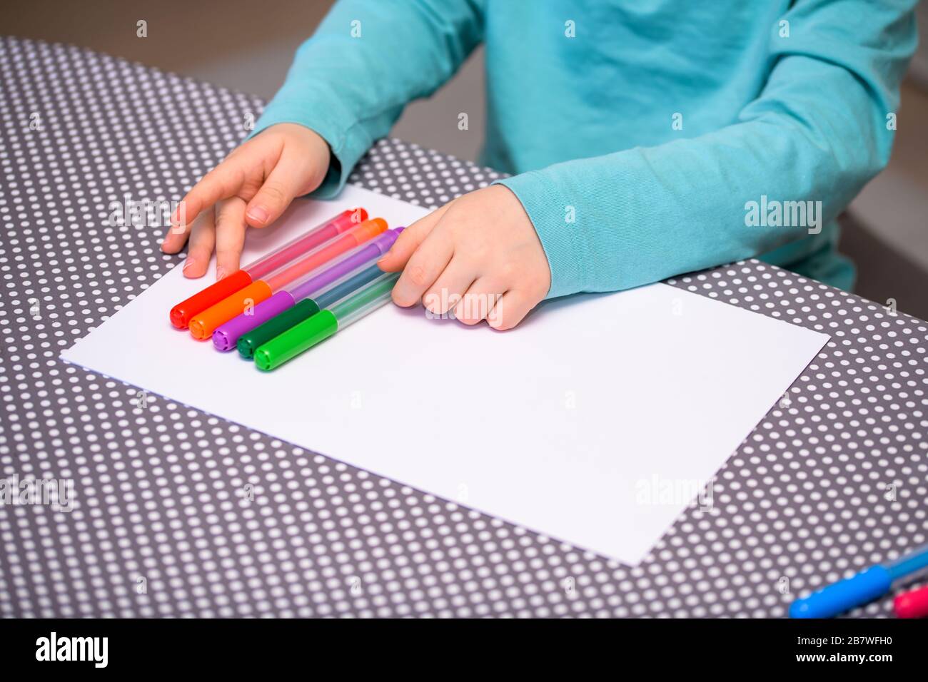 Primo piano di un ragazzo di cinque anni che gioca e allineano le penne a colori su una carta bianca posta su un tavolo con puntini bianchi. Foto Stock