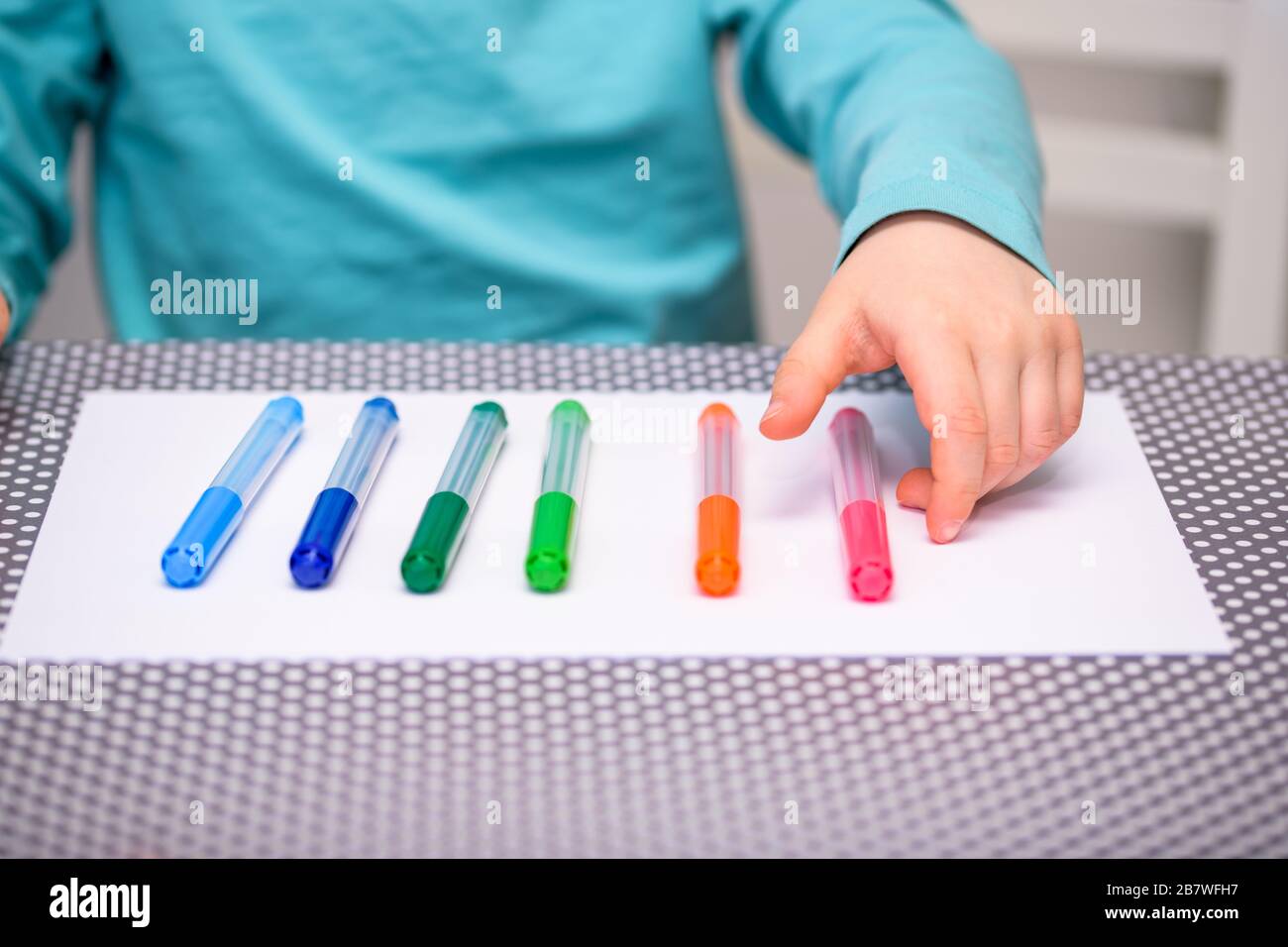 Primo piano di un ragazzo di cinque anni che gioca e allineano le penne a colori su una carta bianca posta su un tavolo con puntini bianchi. Il ragazzo sta raccogliendo una penna rosa. Foto Stock