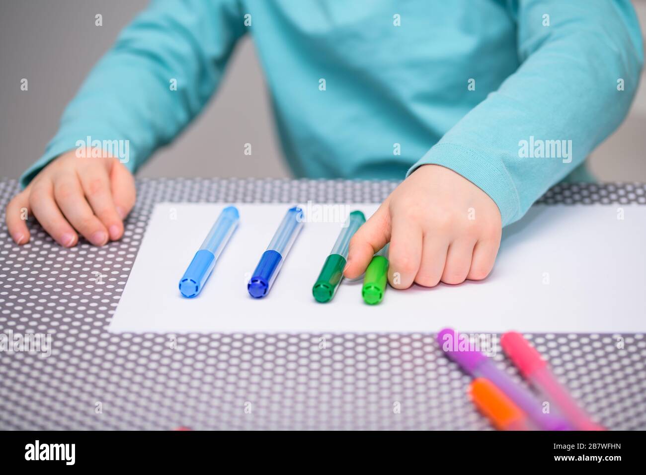 Primo piano di un ragazzo di cinque anni che gioca e allineano le penne a colori su una carta bianca posta su un tavolo con puntini bianchi. Il ragazzo sta tenendo una penna verde. Foto Stock