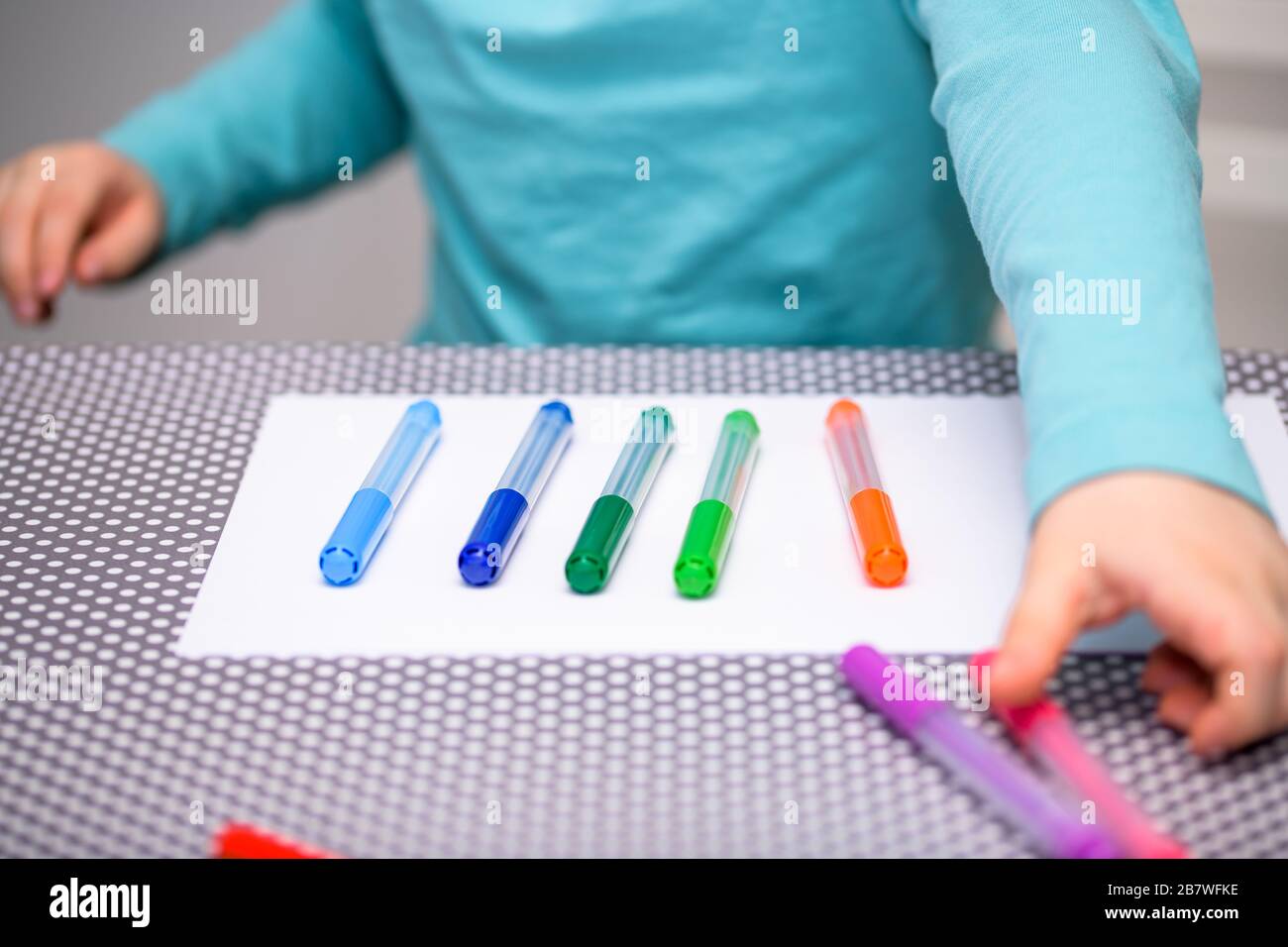 Primo piano di un ragazzo di cinque anni che gioca e allineano le penne a colori su una carta bianca posta su un tavolo con puntini bianchi. Il ragazzo sta raccogliendo una penna rosa. Foto Stock