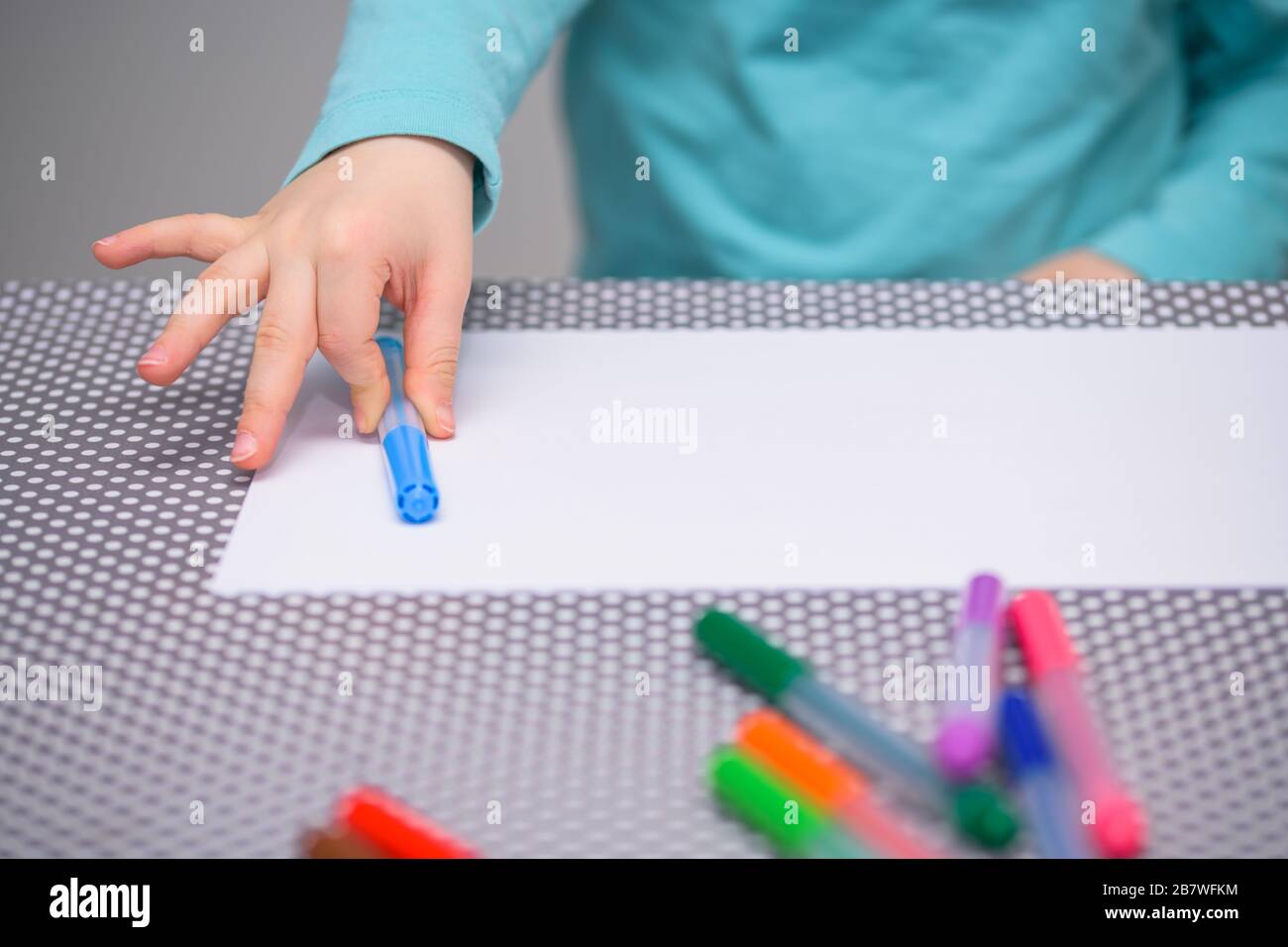 Primo piano di un ragazzo di cinque anni che mette una penna blu chiaro su una carta bianca sopra un tavolo con puntini bianchi. Il ragazzo è vestito con una camicia turchese. Foto Stock