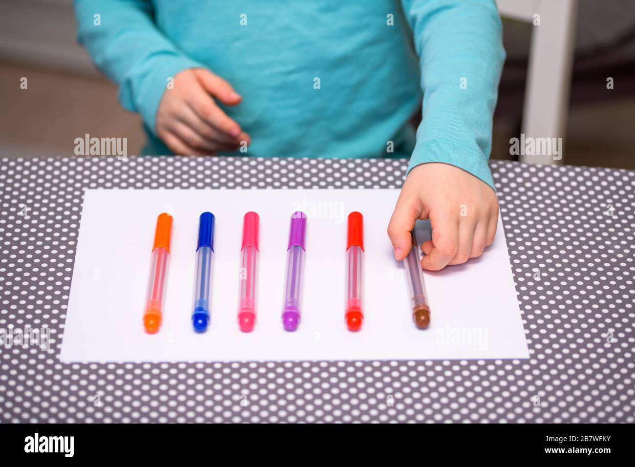 Primo piano di un ragazzo di cinque anni che gioca e allineano le penne a colori su una carta bianca posta su un tavolo con puntini bianchi. Il ragazzo sta tenendo una penna marrone. Foto Stock