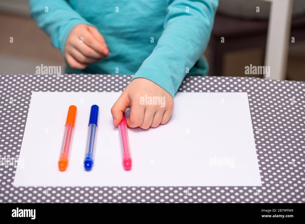 Primo piano di un ragazzo di cinque anni che gioca e allineano le penne a colori su una carta bianca posta su un tavolo con puntini bianchi. Il ragazzo sta tenendo una penna rosa. Foto Stock