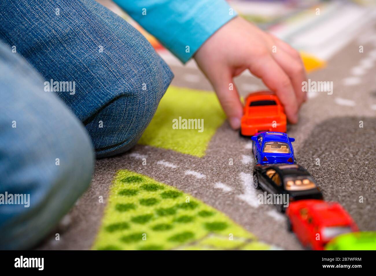 Primo piano di un ragazzo di cinque anni che gioca e allineano le auto giocattolo su un tappetino da gioco con le strade. Il ragazzo è vestito di jeans blu e tiene una macchina giocattolo rossa. Foto Stock