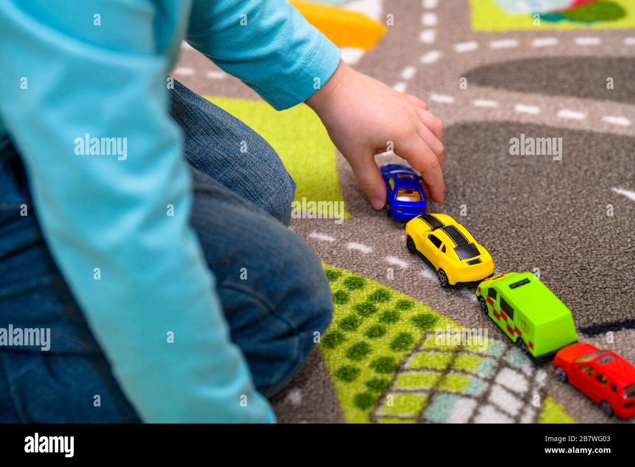 Primo piano di un ragazzo di cinque anni che gioca e allineano le auto giocattolo su un tappetino da gioco con le strade. Il ragazzo è vestito di jeans blu e tiene una macchina blu. Foto Stock
