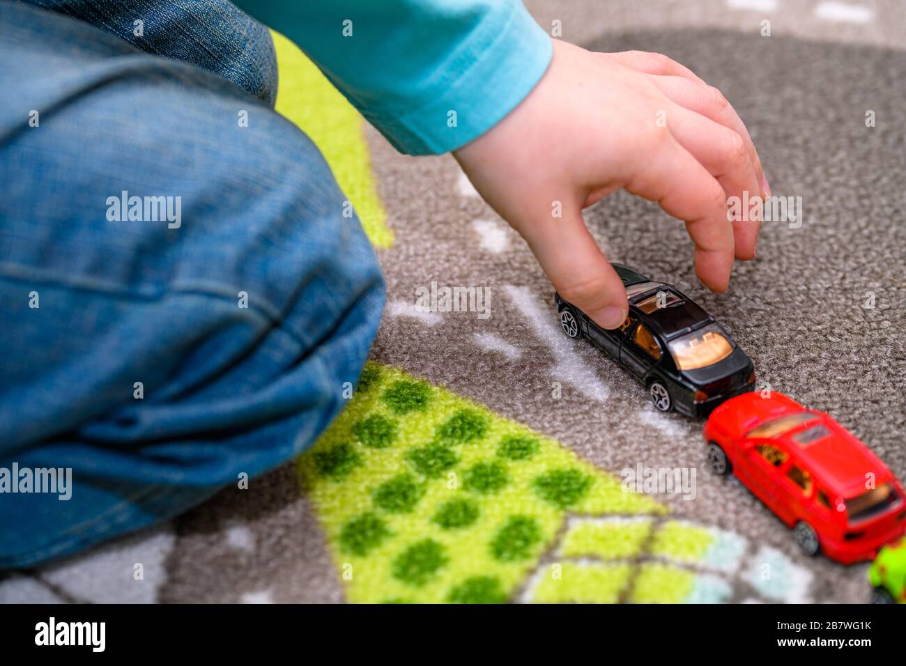 Primo piano di un ragazzo di cinque anni che gioca e allineano le auto giocattolo su un tappetino da gioco con le strade. Il ragazzo è vestito con jeans blu e raccoglie una macchina nera. Foto Stock