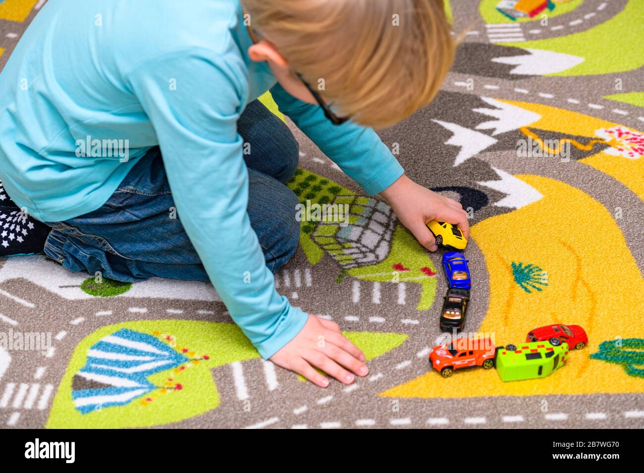Ragazzo di cinque anni che gioca e allineano le auto giocattolo su un tappetino da gioco con le strade. Il ragazzo è vestito di jeans blu e tiene una macchina gialla. Foto Stock