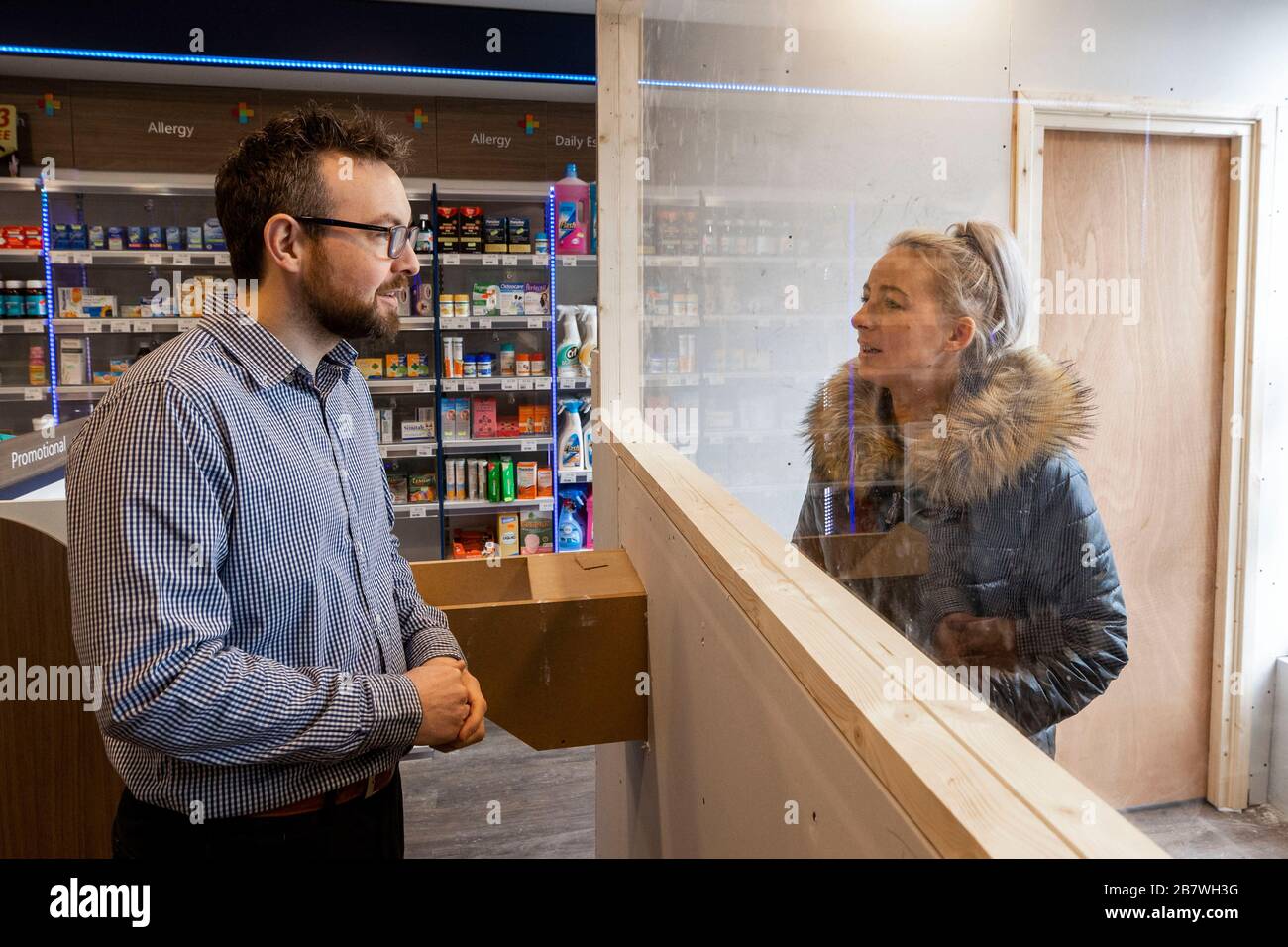 Peter Rice, Group Manager della McKenzie's Pharmacy on the Falls Road a Belfast, parlando con il cliente Laura Ryan utilizzando il loro banco appena installato. Il banco in stile ufficio postale è stato costruito per proteggere il personale e i clienti da Coronavirus. Foto Stock