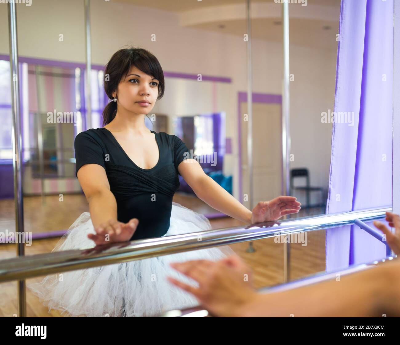 Giovane donna bruna sottile in tuta sportiva nera e tutu bianco fare classe di balletto vicino macchina balletto e guardando in specchio in studio di danza. Moder Foto Stock