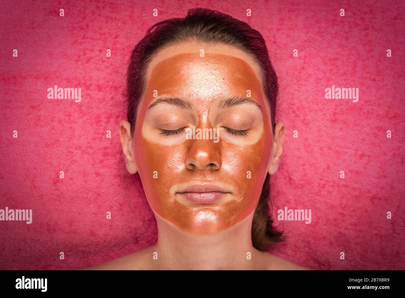 Bella donna con una maschera di bronzo sulla sua faccia. Concetto di cura della pelle e spa. Sfondo corallo Foto Stock