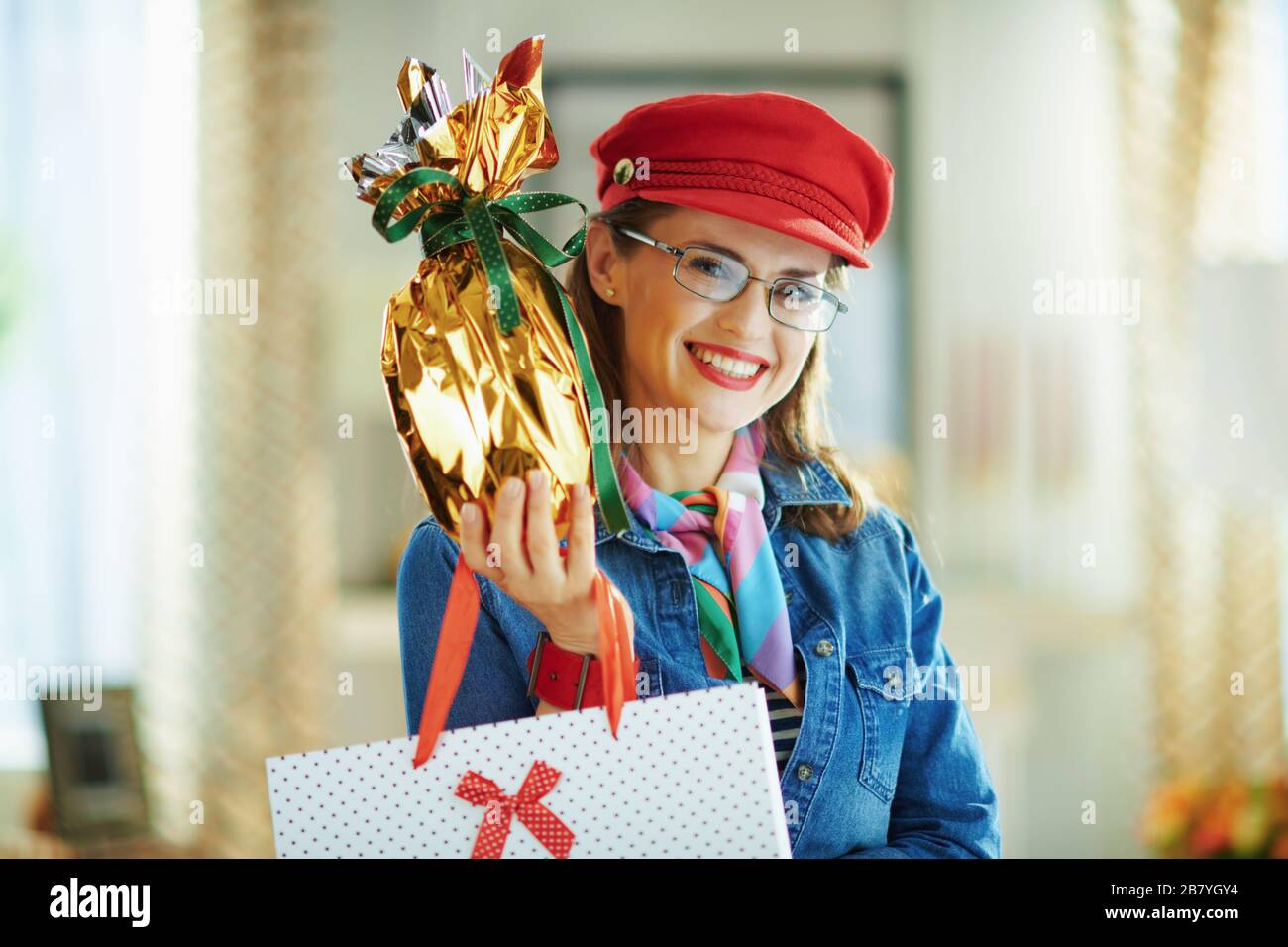 felice donna moderna in una camicia jeans e cappello rosso in occhiali con avvolto in foglio d'oro grande uovo di pasqua e bianco polka puntino shopping bag nel hou moderno Foto Stock