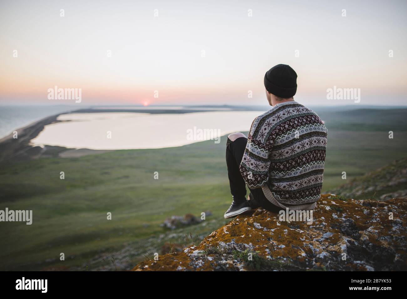 Giovane uomo in maglione a righe seduto in collina al tramonto Foto Stock