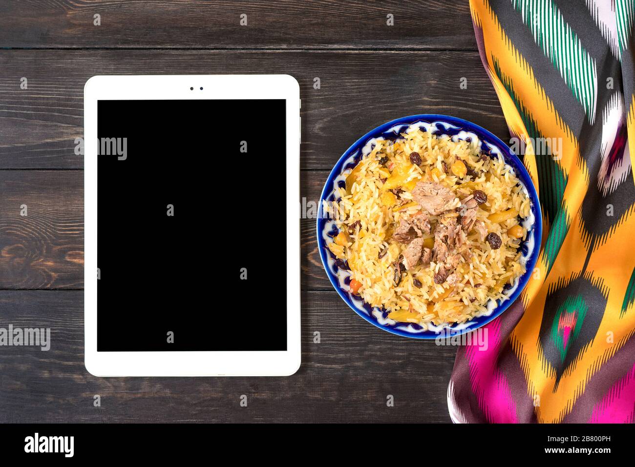 Pilaf Uzbeco nazionale con carne in piastra con modello tradizionale, tessuto ikat, tablet su tavola in legno Vista dall'alto Online food order, concetto di consegna FLA Foto Stock