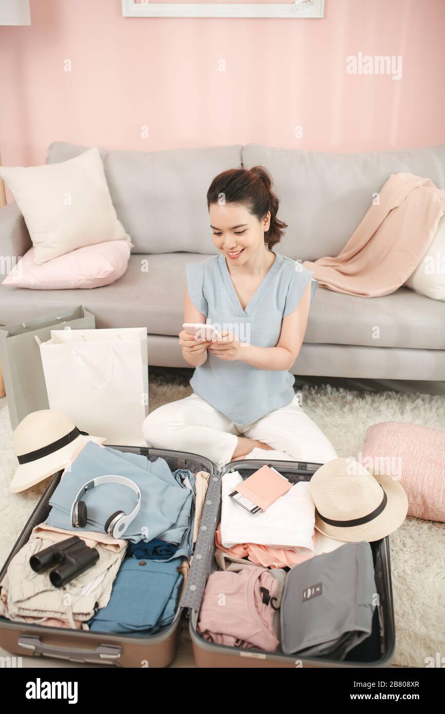 Vista superiore di una donna sorridente ed elegante a casa moderna nella soleggiata giornata estiva utilizzando l'app mobile agente di viaggio sullo smartphone vicino alla valigia da viaggio aperta. Foto Stock