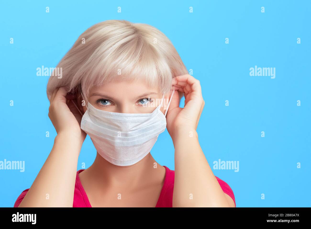 Giovane bella ragazza con capelli biondi mette su una maschera medica protettiva sul suo viso. Protezione dai virus, concetto di prevenzione dei virus su sfondo blu con Foto Stock