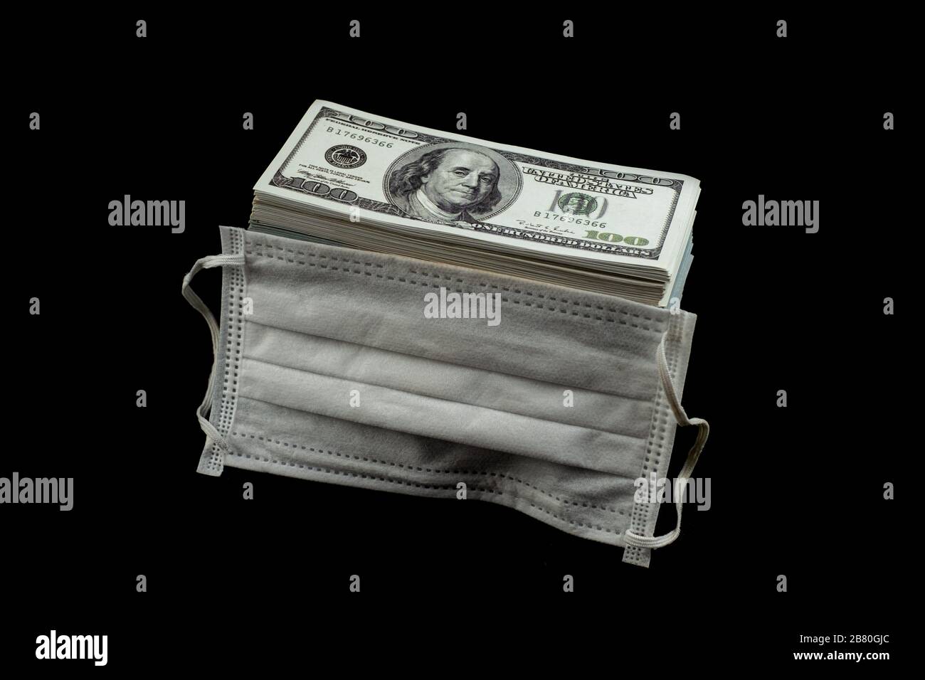 Maschera medica e protezione antivirus e denaro una pila di banconote da 100 dollari un sacco di su sfondo nero, che era costato caro prezzo e. Foto Stock