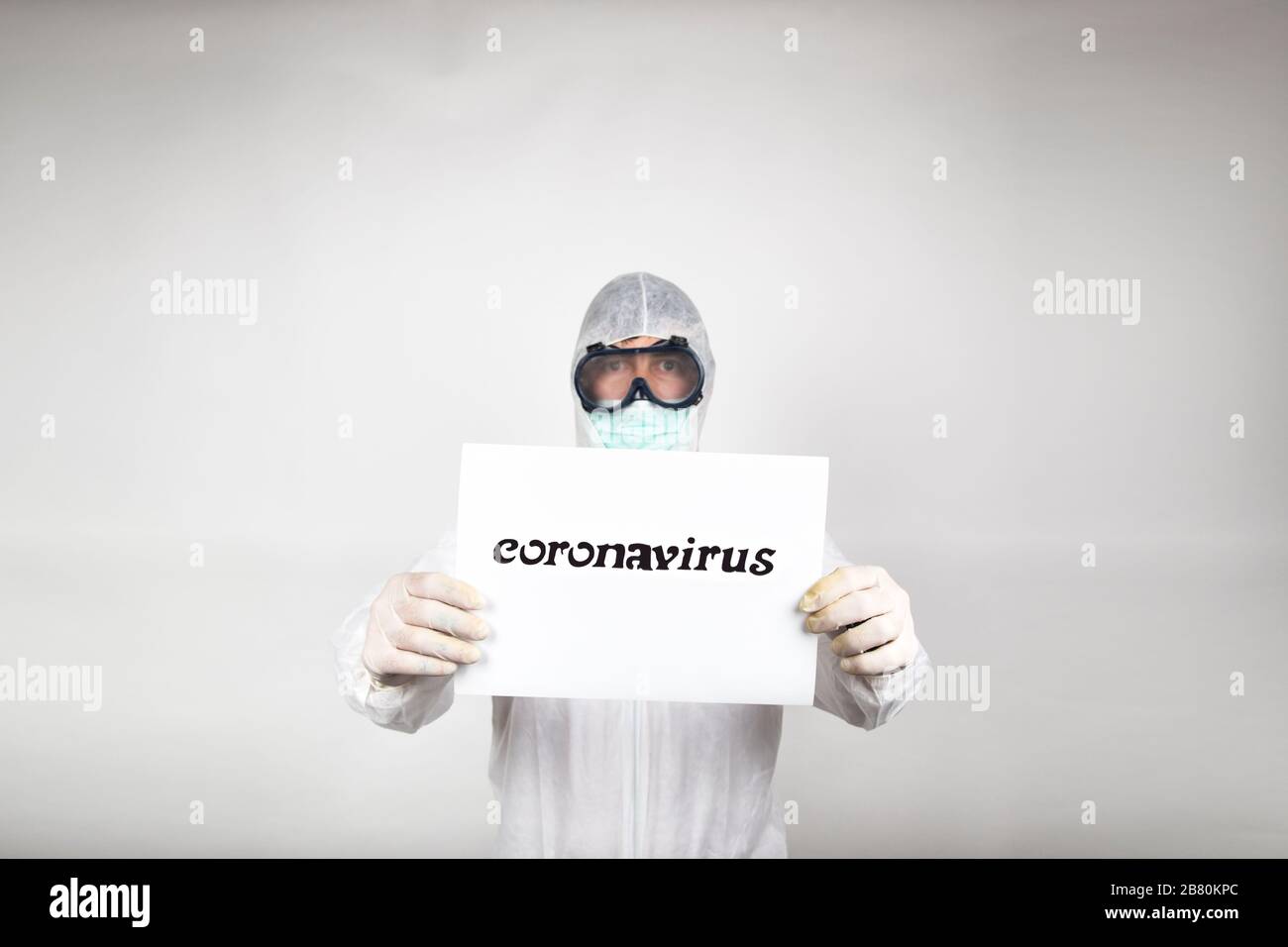 Ritratto dell'uomo in maschera chirurgica con tuta protettiva bianca e un segno con la parola coronavirus isolato in studio su sfondo bianco. Prevenzione Foto Stock