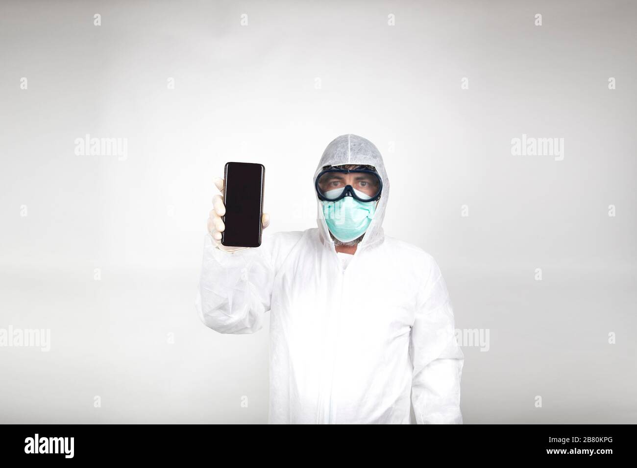 Ritratto dell'uomo in maschera chirurgica indossando tuta protettiva bianca con telefono cellulare isolato in studio su sfondo bianco. Prevenzione contro coronaviru Foto Stock